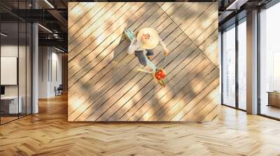 Above and diagonal shot of a woman riding a bicycle on a wooden pier carying tomatoes and wearing a straw hat . There is afternoon sun and shadows on the pier. Wall mural