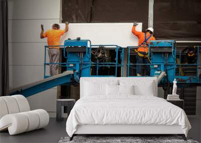 Two construction workers wearing hardhats on a blue manlift hanging section of drywall paneling on prefab building Wall mural