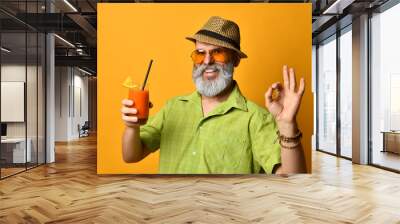 Bearded grandpa in hat, green shirt, sunglasses. Smiling, showing okay sign, holding glass of juice, posing on orange background Wall mural