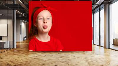 Cute funny red-haired girl in a red bandage on her hair and a red T-shirt jokingly shows your tongue. Wall mural