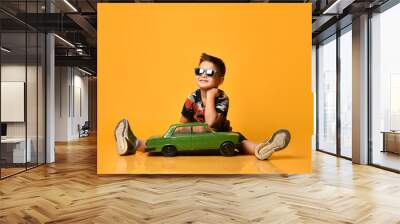 Kid in sunglasses, camouflage t-shirt, sneakers. Sitting on floor, holding green model of retro car, posing on orange background Wall mural