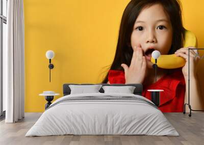 Little asian kid in red blouse. She pretending to be talking on banana like by phone, posing on orange background. Close up Wall mural