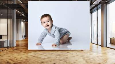 Little baby boy toddler in grey casual jumpsuit and barefoot crawling on floor, smiling and looking up over white wall background Wall mural