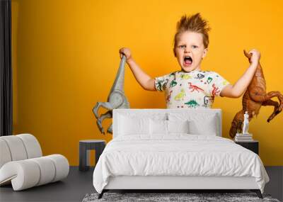 little cheerful boy holds a plastic dinosaur in each hand. The boy is dressed in a suit with pictures of dinosaurs. Wall mural