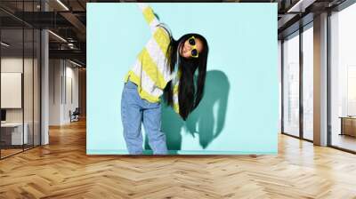 Portrait of stylish bright Korean girl posing over blue background with outstretched hands. A child in a bright sweater, jeans, sneakers and sunglasses. Kids fashion and style concept. Wall mural