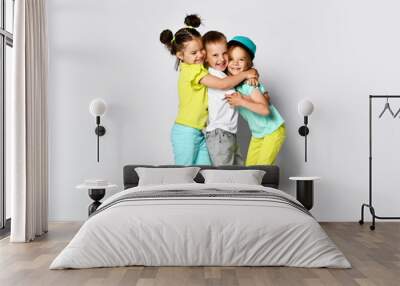 Studio portrait of children on a light background: full body shot of three children in bright clothes, two girls and one boy. Triplets, brother and sisters. hugging on camera. Family ties, friendship Wall mural