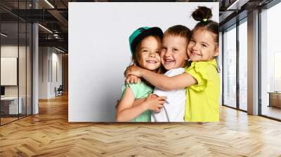 Studio portrait of children on a light background: full body shot of three children in bright clothes, two girls and one boy. Triplets, brother and sisters. hugging on camera. Family ties, friendship Wall mural