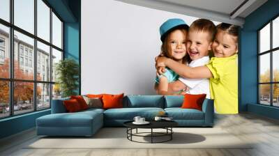 Studio portrait of children on a light background: full body shot of three children in bright clothes, two girls and one boy. Triplets, brother and sisters. hugging on camera. Family ties, friendship Wall mural