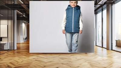 Studio shot of a little girl in warm outerwear, blue sleeveless vest, jeans and sweatshirt, walking towards the camera, isolated on white background. Children's fashion clothing and fashion Wall mural