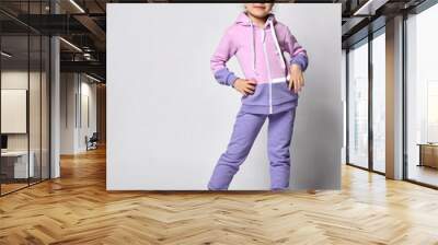 Studio shot of a small blonde girl in pink violet tracksuit with a hood, posing for the camera with her hand, staring at the camera, isolated on white background. Wall mural