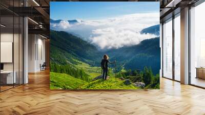 Woman resting on mountain top, looking at view dramatic landscape clouds over the valley, clear blue sky. Summer activity fitness wellbeing freedom success. Wall mural