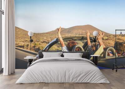 Back view group three euphoric friends in convertible car twisting and waving Two curly girls and charming bearded man on vacation having fun in topless auto Arms up palms open driving in the sunlight Wall mural