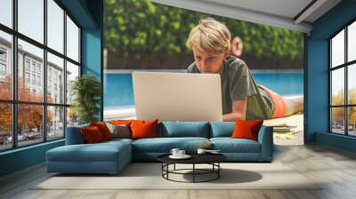 Beautiful young boy using laptop near the swimming pool in a sunny summer day Child doing homeworks outdoors after the end of school. Technology allows to stay connected with remote friends everywhere Wall mural