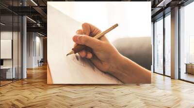 Close up view of a young hand drawing on a white sheet. Kid hold a black wooden pencil and draw something on a warm orange light at home. Children writing on a paper. Teen drawing freehand a manga. Wall mural