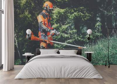 Man holding a brushcutter cut grass and brush. Lumberjack at work wears orange personal protective equipment. Gardener working outdoor in the forest. Security, occupation, forestry, worker, concept Wall mural