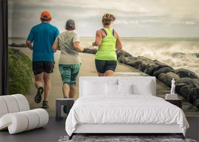 Three people running along the ocean. Two girls and one man jogging together on a beautiful road along the sea with big waves and spray. A beautiful day of outdoors sport in New Zealand. Wall mural