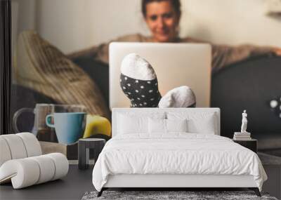 Woman sitting on a couch in the living room with warm socks in a winter morning. Girl using laptop and works at home, having natural breakfast with tea coffee and fruits. Focus on feet in foreground Wall mural