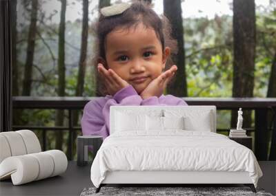 An Asian little girl with curly hair in purple long sleeve shirt posing adorable with trees or nature background Wall mural