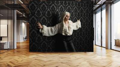 close up portrait of beautiful blonde female model, wearing vintage gothic fantasy ruffle blouse white shirt, leather pants and long blonde plaited hair.  moody black brocade wall paper background Wall mural