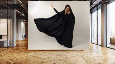 full length portrait of a blonde lady wearing black lace dress and hooded cloak,  standing pose, isolated against creamy studio background. Wall mural