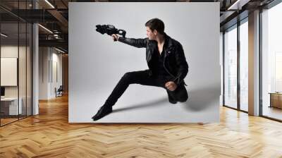 Full length portrait of a  brunette man wearing leather jacket  and holding a science fiction gun.  kneeling  action pose isolated  against a grey studio background.
 Wall mural