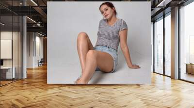 full length  portrait of girl wearing shirt and denim shorts.sitting pose, isolated on grey studio background. Wall mural