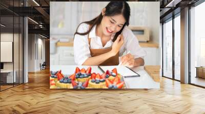 Asian woman bakery shop owner talking on mobile phone with customer about catering bakery order in kitchen. Bakery chef making fruit tart selling online delivery. Small business food and drink concept Wall mural