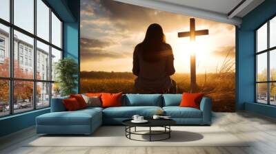 Silhouette of a woman sitting on the grass praying in front of a cross at sunset Wall mural