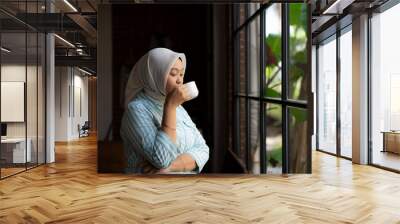 A woman wearing hijab and holding a cup of coffee. A woman wearing a scarf and holding a cup of coffee. She is smiling and looking out the window
 Wall mural