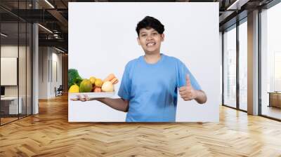 Young excited Indonesian man standing carrying tray of healthy vegetables and fruits with his hand pointing, healthy lifestyle concept, healthy food campaign, isolated on white background. Wall mural