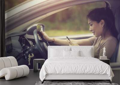 Young woman looking at her smartphone while driving a car on a sunny day with sun light coming through windshield Wall mural