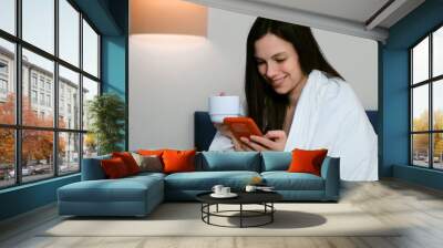 Brunette young smiling joyful woman is swiping pages in smartphone browsing social media wrapped in blanket and drinking her morning coffee sitting in bed. Rest, relax on vacation in bedroom. Wall mural