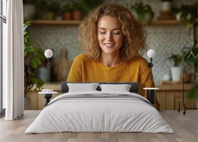 A cheerful woman with curly hair smiles brightly at her laptop, sitting in a modern kitchen with greenery, representing a harmonious work-life balance. Wall mural