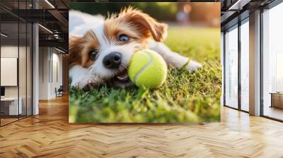 A delightful puppy playfully chewing on a tennis ball, lying on vibrant green grass under the bright sun, evoking a sense of joy and playful energy. Wall mural