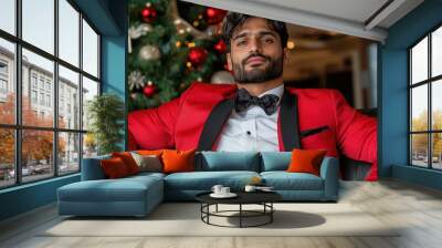 A stylish man sits confidently in a striking red formal suit beside a beautifully decorated Christmas tree, radiating holiday spirit and luxurious festivities. Wall mural