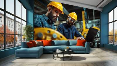 Two engineers in hard hats are intently focused on a laptop amidst mechanical components in a factory, capturing intent concentration and teamwork. Wall mural