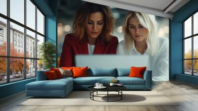 Two women, one in a red blazer and the other in a white blouse, are focused on working together on a laptop, representing teamwork and collaboration in a modern office setting. Wall mural