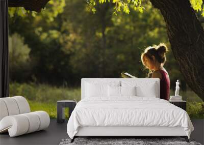 sunny portrait of a beautiful girl sitting on green glade under tree branches with book, woman reading novel on nature, concept hobby and lifestyle Wall mural