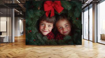 Two happy kids hiding behind the Christmas wreath, their happy faces peeking out. A boy and a girl, with a red bow on top of the wreath and green pine branches surrounding them. Wall mural