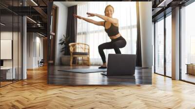 A beautiful young asian woman doing squats while watching online workout tutorials on laptop at home Wall mural