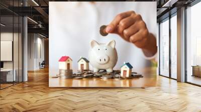 A woman putting coins into piggy bank with house models on pile of coins for saving money concept Wall mural