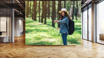 An asian female traveler with a hat and backpack looking at a watch in a beautiful pine woods Wall mural