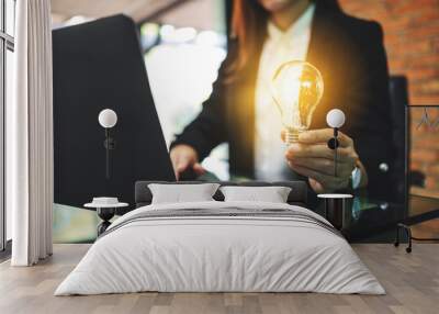 Closeup image of a businesswoman holding a glowing light bulb while working on laptop computer in office Wall mural