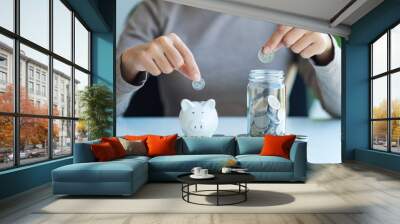 Closeup image of a woman putting coin into piggy bank and a glass jar for saving money concept Wall mural