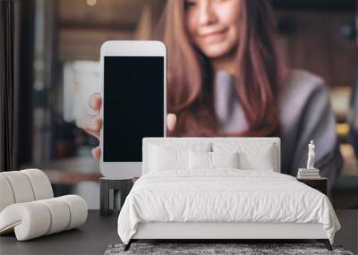 Mockup image of a beautiful asian woman holding and showing white mobile phone with blank black desktop screen in cafe Wall mural