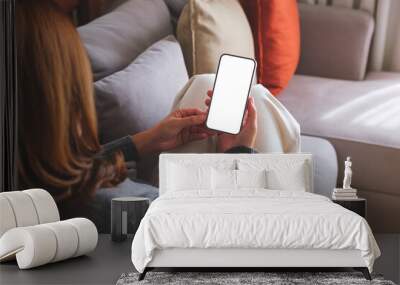 Mockup image of a woman holding mobile phone with blank desktop white screen while lying on a sofa at home Wall mural