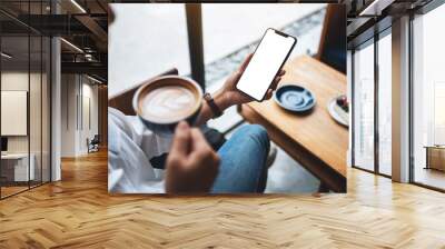 Mockup image of a woman holding mobile phone with blank white desktop screen while drinking coffee in cafe Wall mural