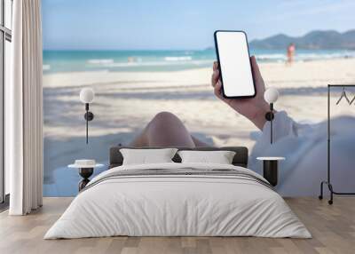 Mockup image of a woman holding white mobile phone with blank desktop screen while laying down on beach chair on the beach Wall mural