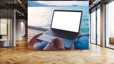 Mockup image of a woman using laptop with blank white desktop screen while sitting by the sea with blue sky background Wall mural