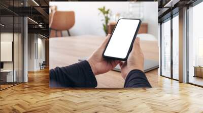 Mockup image of hands holding black mobile phone with blank white screen with laptop on wooden table Wall mural